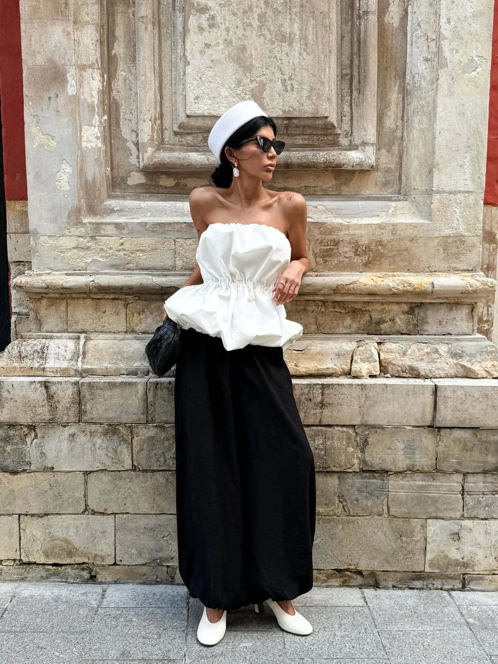 Woman in a white strapless top and black wide-leg pants standing against a stone wall.
