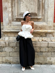 Woman in a white strapless top and black wide-leg pants standing against a stone wall.