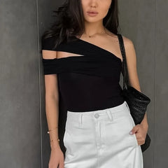 Woman wearing a black one-shoulder top and white pants against a gray wall.