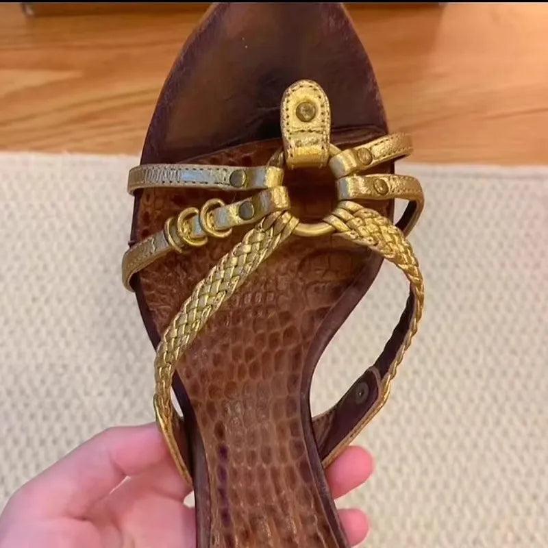Brown sandal with gold straps held by a hand on a wooden floor.