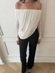 Person wearing a white off-shoulder top and black pants against a white wall.