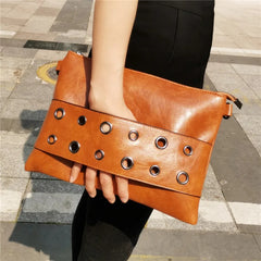 Brown leather clutch with grommet details held by a person on a pavement.