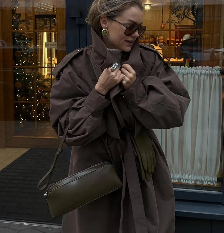 Woman in a brown coat and sunglasses holding a green handbag, standing in front of a store window.