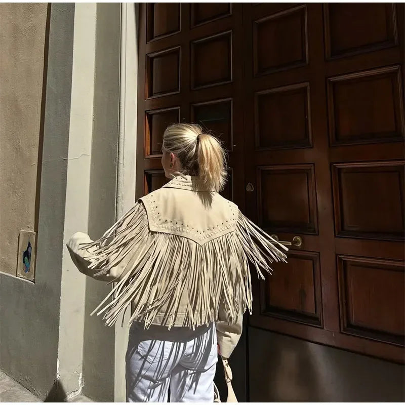 Person wearing a beige fringed jacket standing in front of a large wooden door.