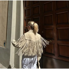 Person wearing a beige fringed jacket standing in front of a large wooden door.