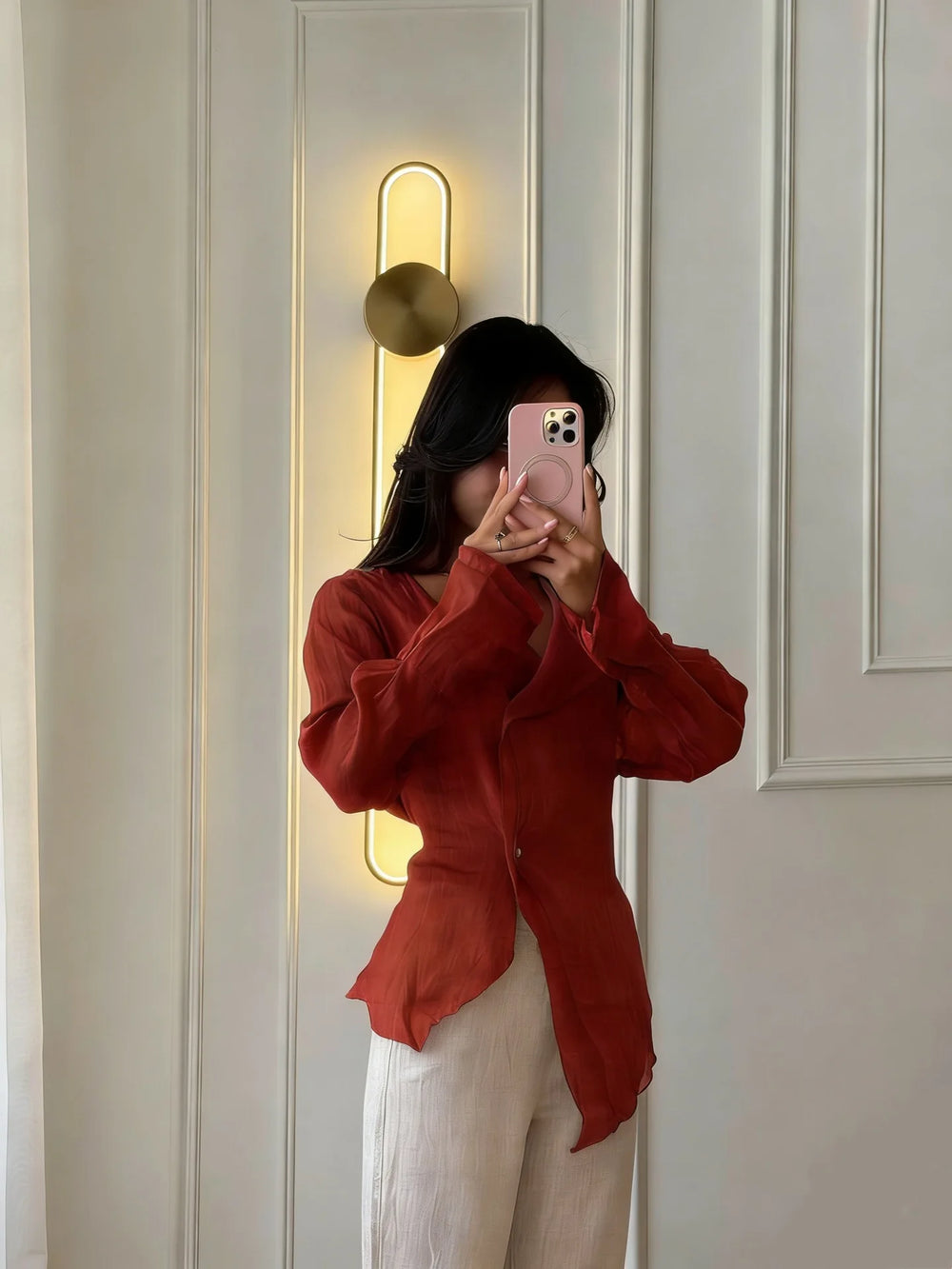 Person wearing a red sweater taking a mirror selfie in a room with white walls and a gold light fixture.