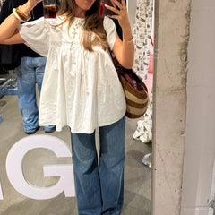 Person taking a mirror selfie wearing a white blouse and blue jeans, holding drinks.