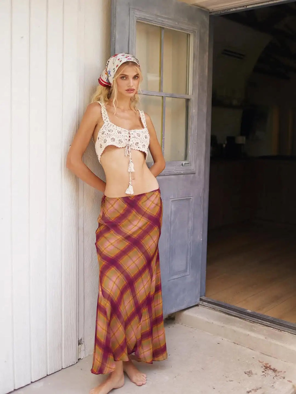 Woman leaning against a door wearing an irregular hem plaid maxi skirt and a crochet top.