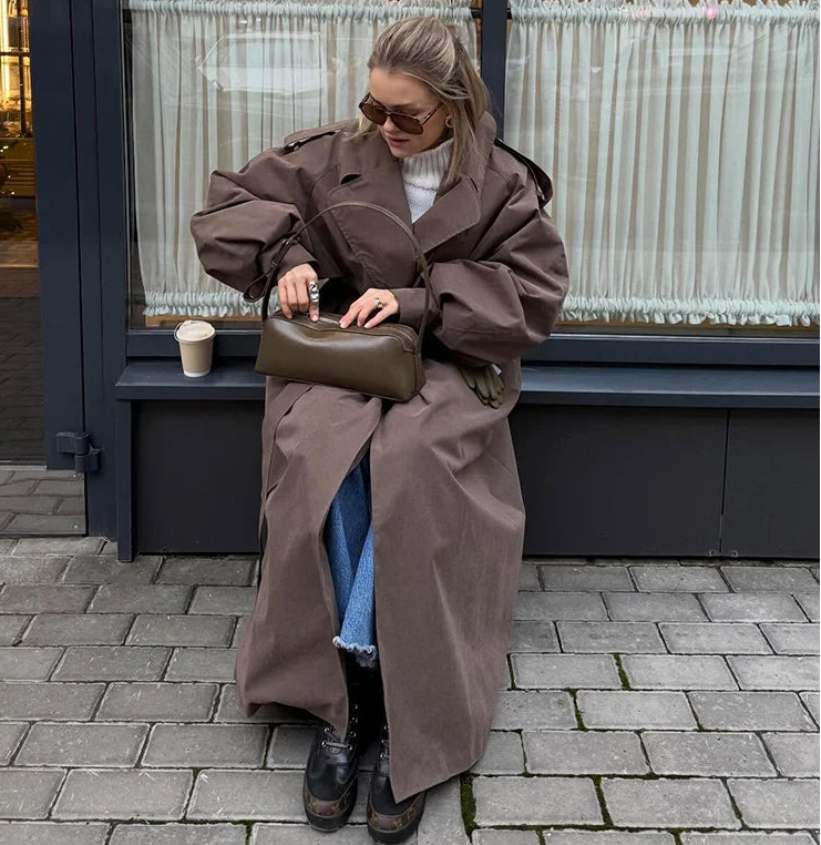 Person wearing a long brown coat sitting on a bench with a handbag.