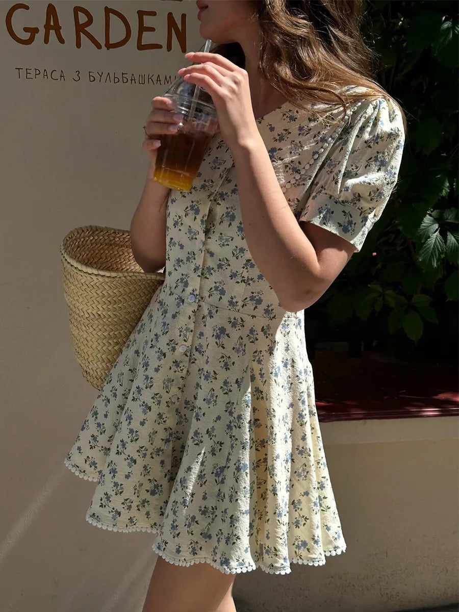 Woman in a floral dress holding a drink, standing in a garden setting.