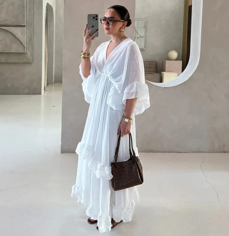 Woman in a white dress holding a brown handbag, standing in a minimalistic room.