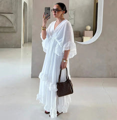 Woman in a white dress holding a brown handbag, standing in a minimalistic room.