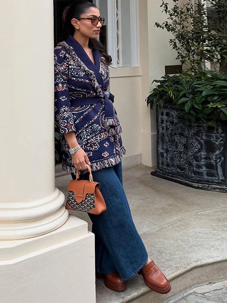 Woman in a patterned blazer and blue jeans holding a brown handbag, standing on a sidewalk.