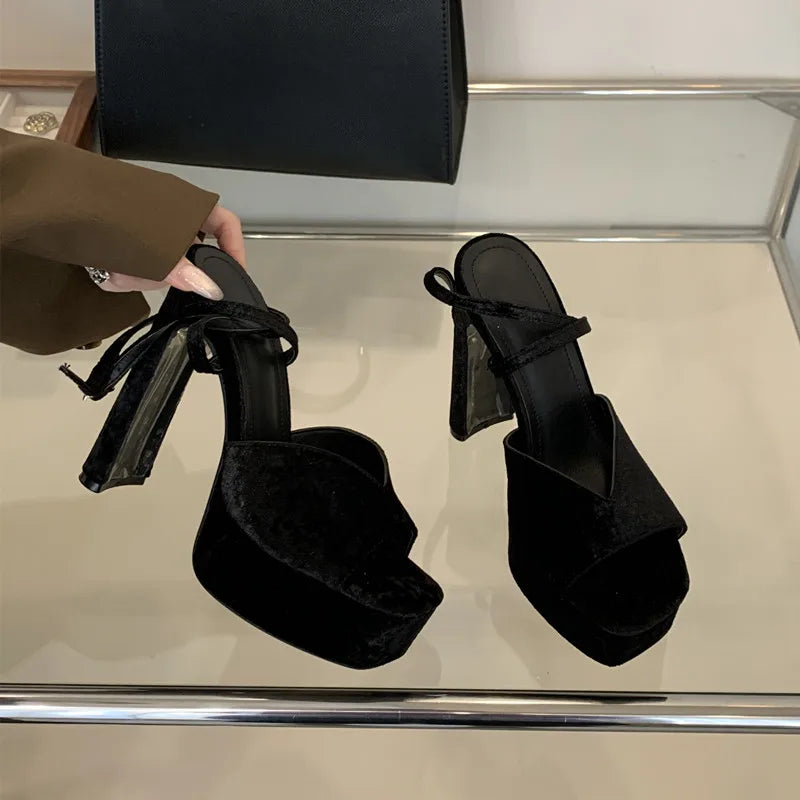 Black high-heeled sandals with ribbon details on a reflective surface.