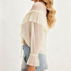 Woman wearing a sheer beige top with ruffled details and denim shorts on a white background