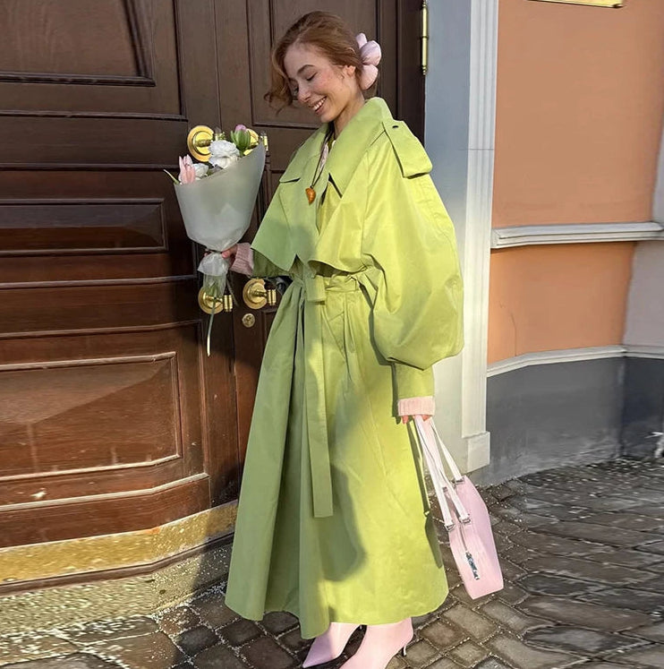 Woman in a bright green coat holding flowers and a pink bag on a street.