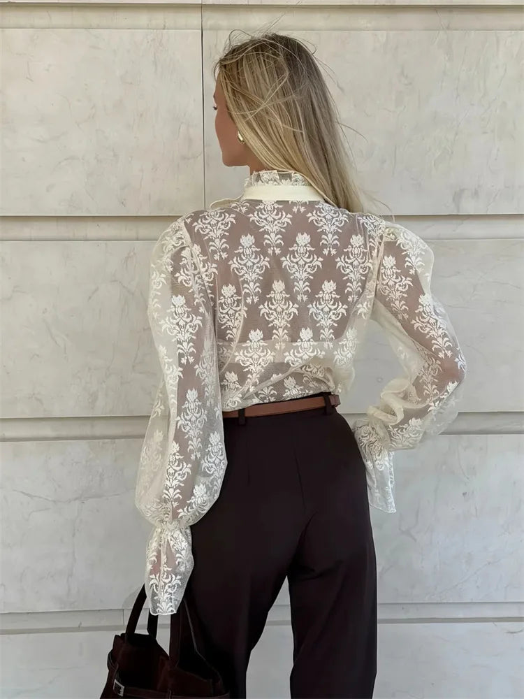 Person wearing a sheer lace blouse with long sleeves against a light-colored wall.