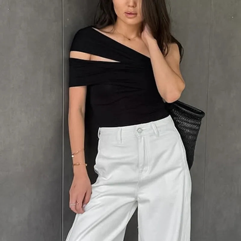 Woman wearing a black one-shoulder top and white pants against a gray wall.