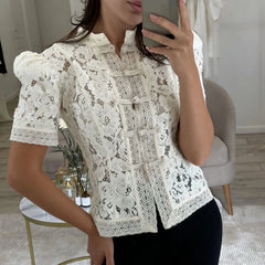 Person wearing a white lace blouse in a room with a chair and table.
