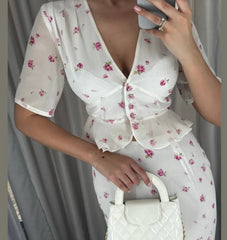 Woman wearing a white floral dress holding a white handbag against a gray curtain background