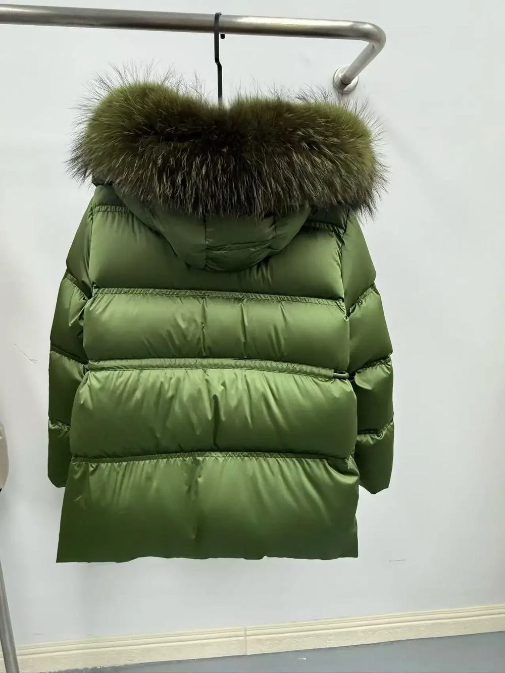 Green puffer jacket with fur hood on a hanger against a white wall.