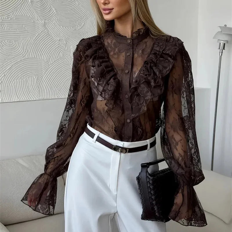 Woman wearing a sheer brown blouse with ruffles and white pants in an indoor setting.