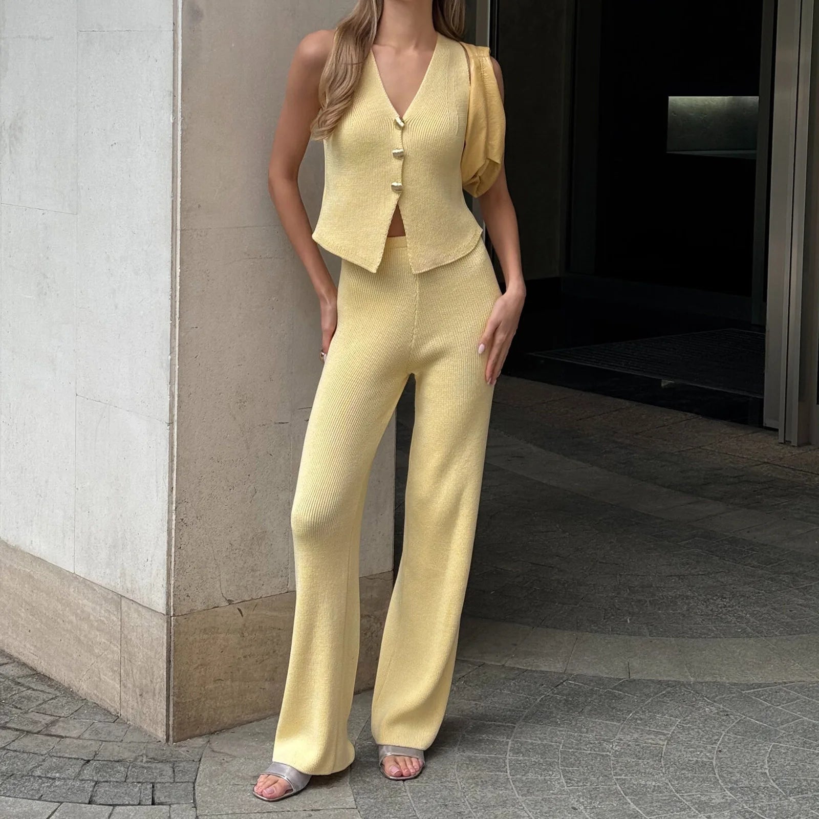 Woman wearing a sleeveless yellow vest and knit trousers standing at a modern building entrance
