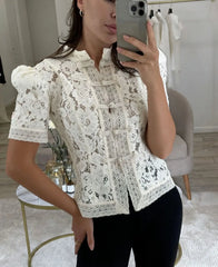 Woman taking a mirror selfie wearing a white lace blouse in a room.