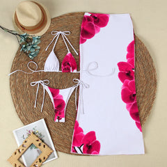 A flat lay of a white bikini top and bottom with pink orchid print, and a matching white maxi skirt with pink orchid print, arranged on a round woven mat.