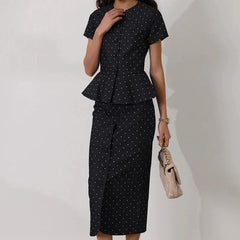 Woman wearing a black polka dot outfit with a peplum top and skirt, holding a beige handbag 