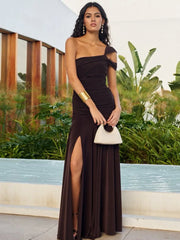 Woman in a black evening gown with a high slit, holding a white clutch, standing outdoors.