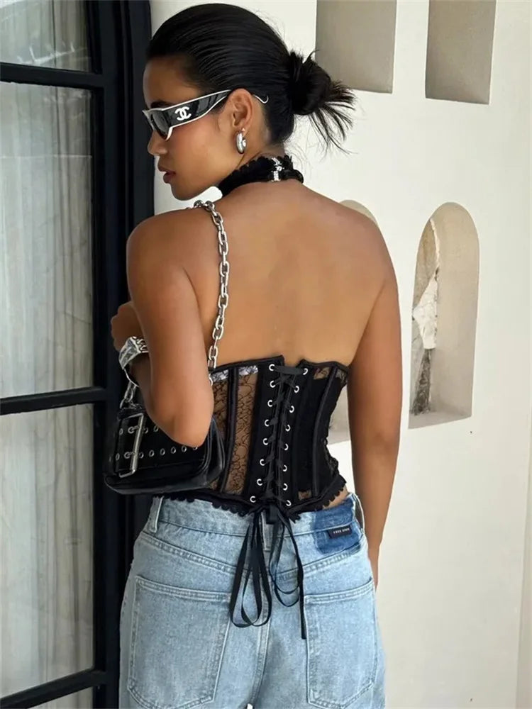 Woman wearing a black lace top and blue jeans, standing against a white wall.