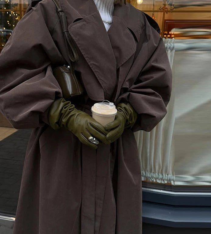 Person wearing a long brown coat and green gloves holding a coffee cup, standing next to a car.