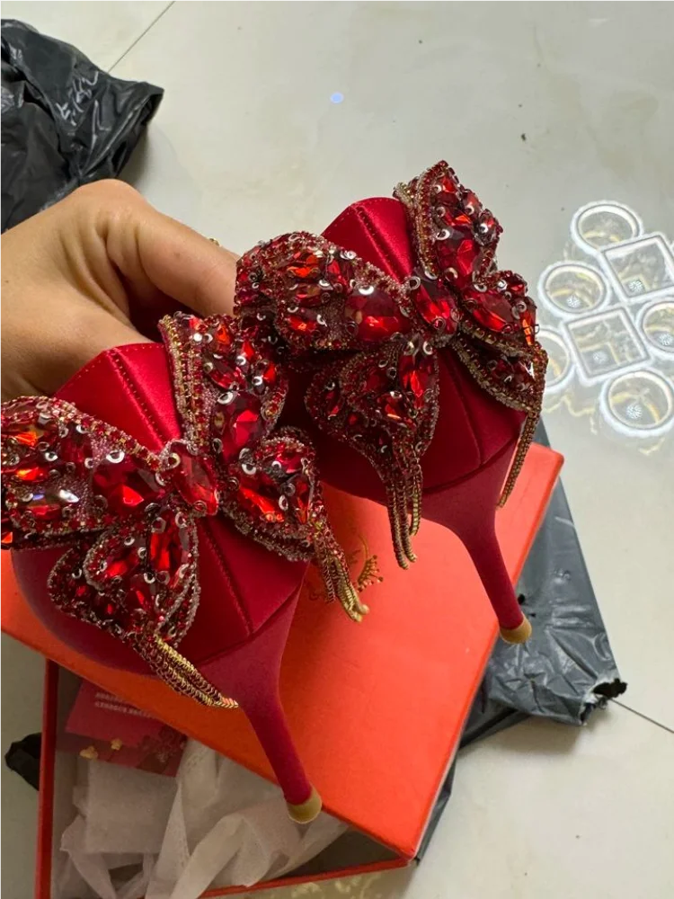 Red high-heeled shoes with decorative elements held by a hand on a light-colored floor.