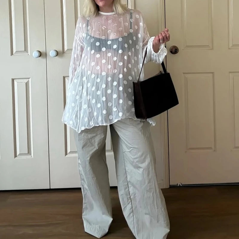 Person wearing a sheer polka dot top and white pants, holding a black handbag indoors.