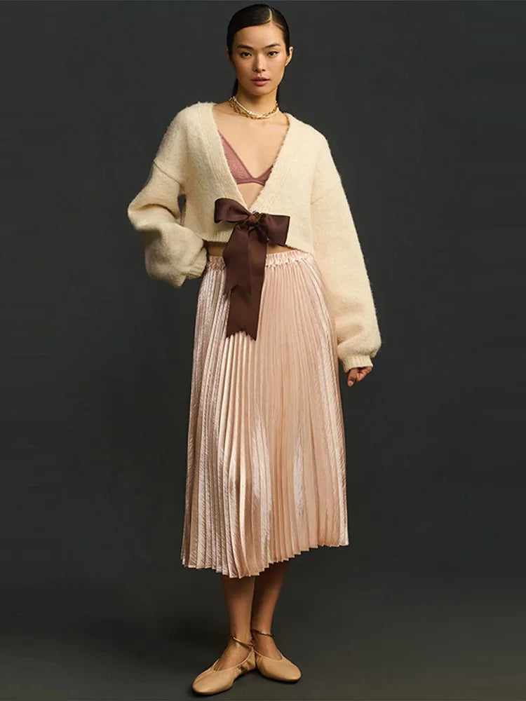 Woman wearing a beige cardigan, pink pleated skirt with a brown bow, and beige shoes on a black background