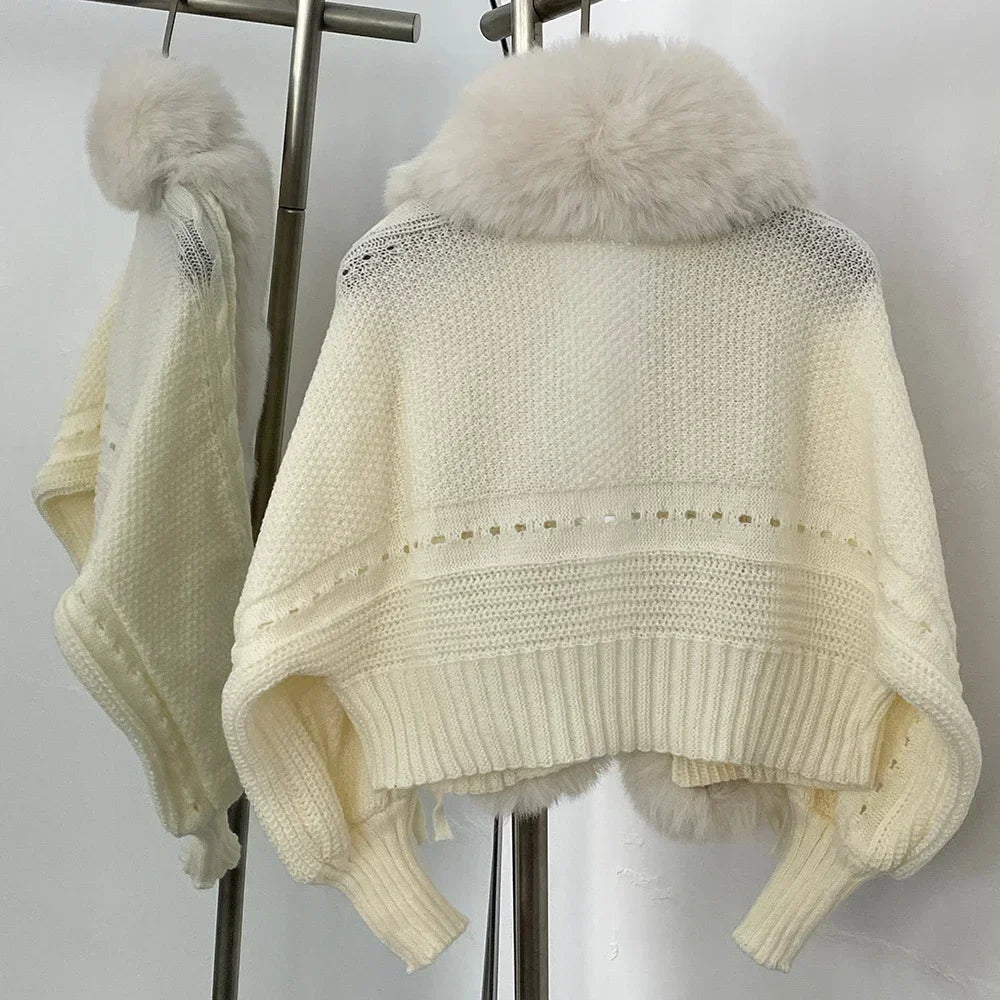 Cream-colored knitted poncho with fur trim on a stand against a white background
