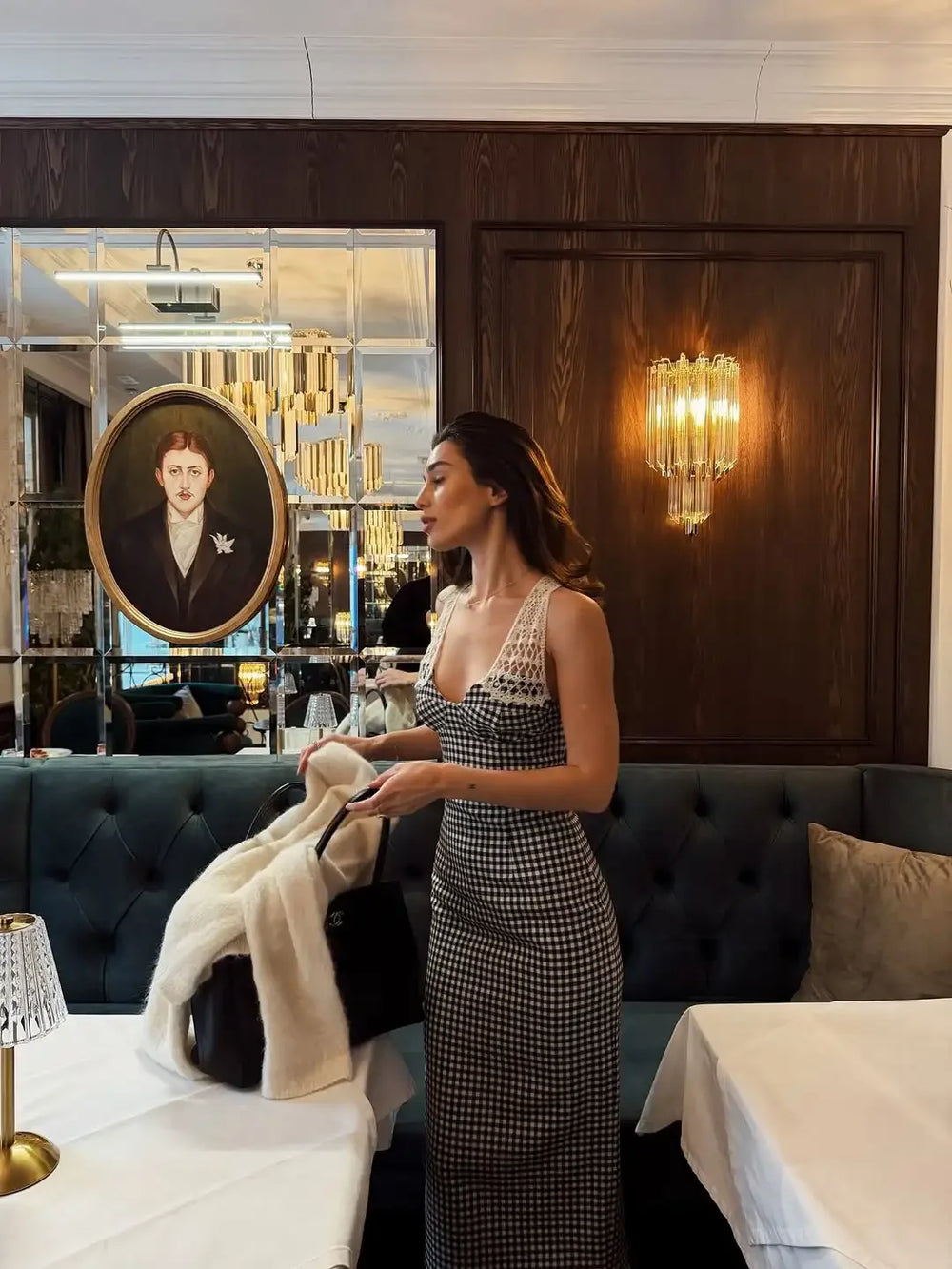 Woman in a patterned dress standing in an elegant interior setting with a portrait and chandelier.