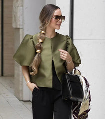 Woman wearing a green top and black pants, standing against a white wall.