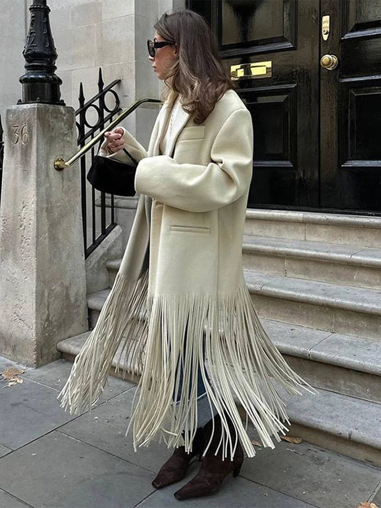 Woman wearing a long cream coat with fringe details, standing on a city street.