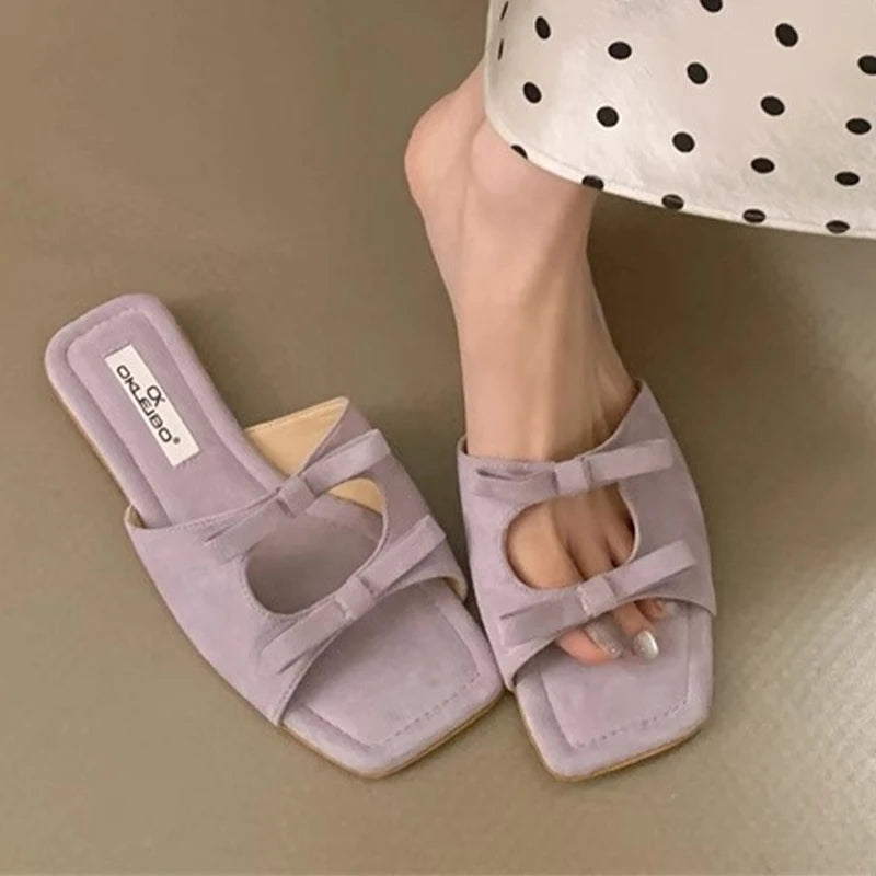 Light purple sandals with a bow detail worn by a person on a beige surface.