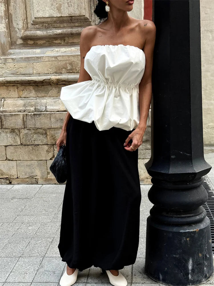 Woman wearing a white strapless top and black skirt standing against a stone wall.