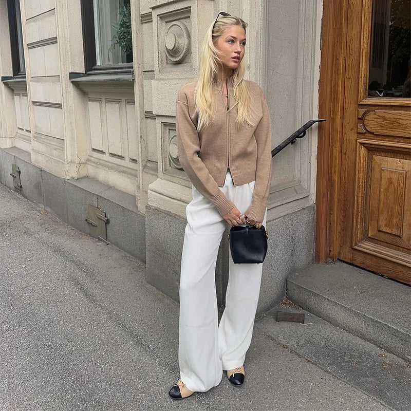 Woman in beige jacket and white pants standing on a street with a building in the background