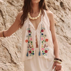 Woman wearing a white sleeveless top with floral embroidery against a stone wall.