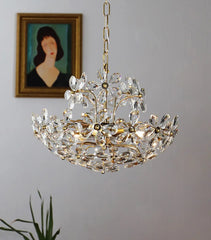 Crystal chandelier hanging against a gray wall with a framed portrait and plant in the background.