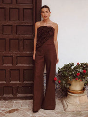 Woman in a brown strapless jumpsuit standing in front of a textured wall with plants.