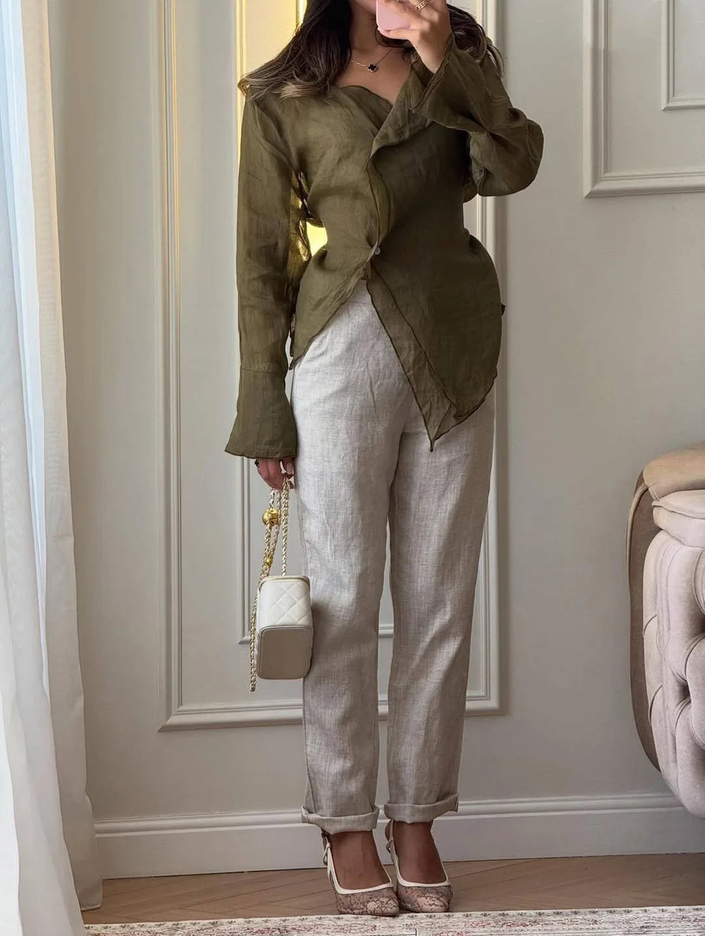 Person wearing a green jacket and beige pants, holding a white handbag indoors.