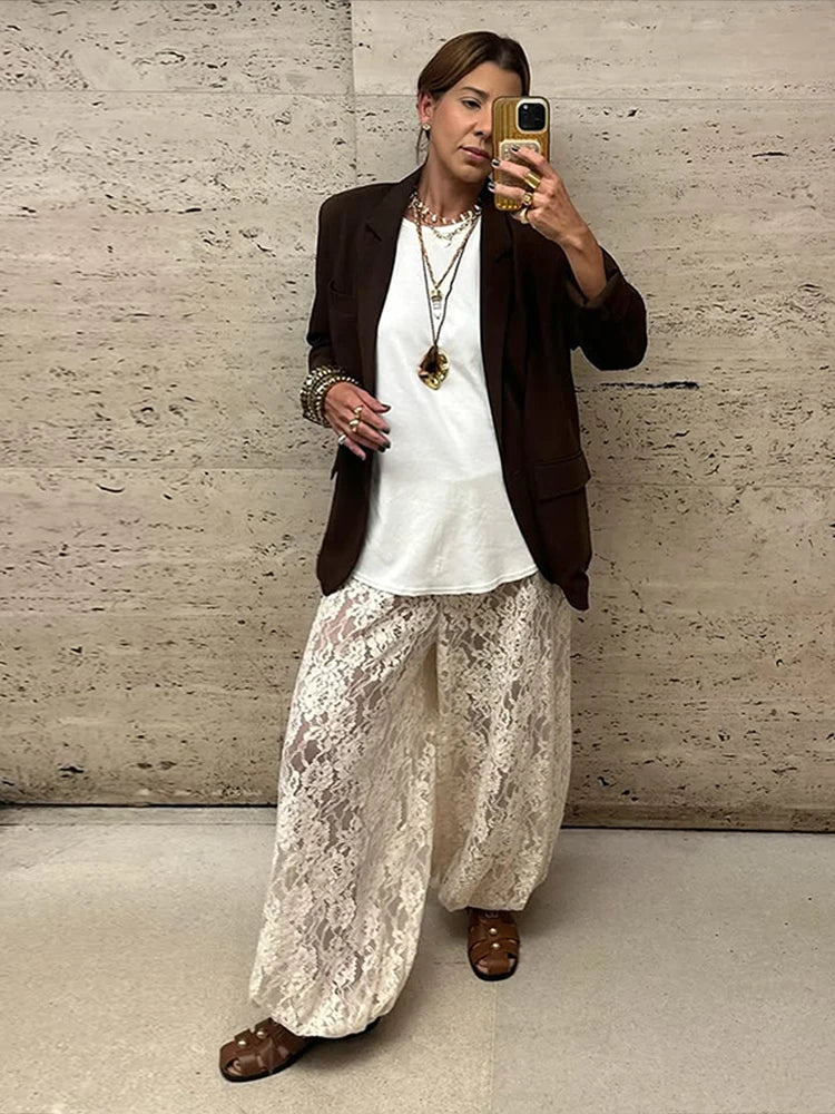 Person taking a mirror selfie wearing a white top, brown blazer, and lace pants against a textured wall.