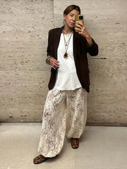 Person taking a mirror selfie wearing a white top, brown blazer, and lace pants against a textured wall.
