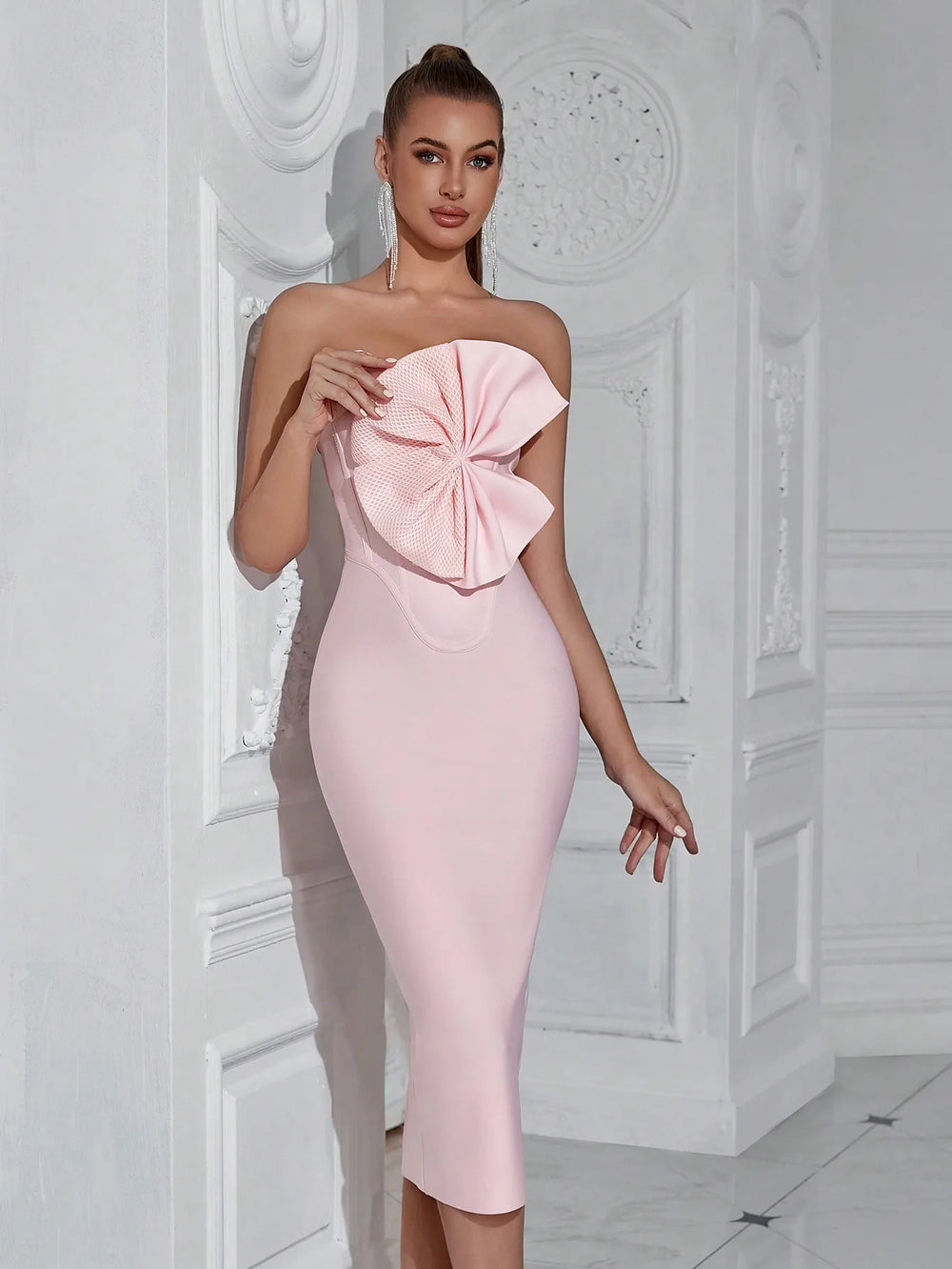 Woman wearing a pink strapless dress with a large bow detail against a white decorative wall.